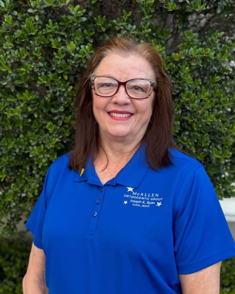 Portrait of Dana Schultz, office manager at McAllen Orthodontic Group in McAllen, TX, wearing a blue clinic polo shirt and standing outdoors in front of greenery.