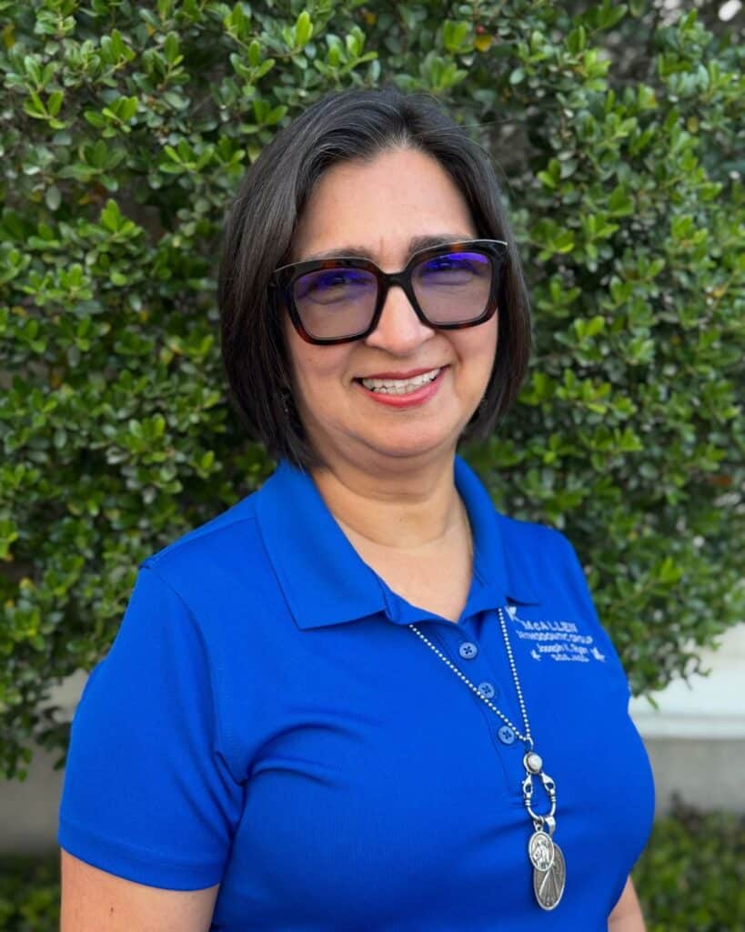 Portrait of Lucy Ortega, team member at McAllen Orthodontic Group in McAllen, TX, wearing a blue clinic polo shirt, glasses, and a necklace while standing outdoors in front of greenery.