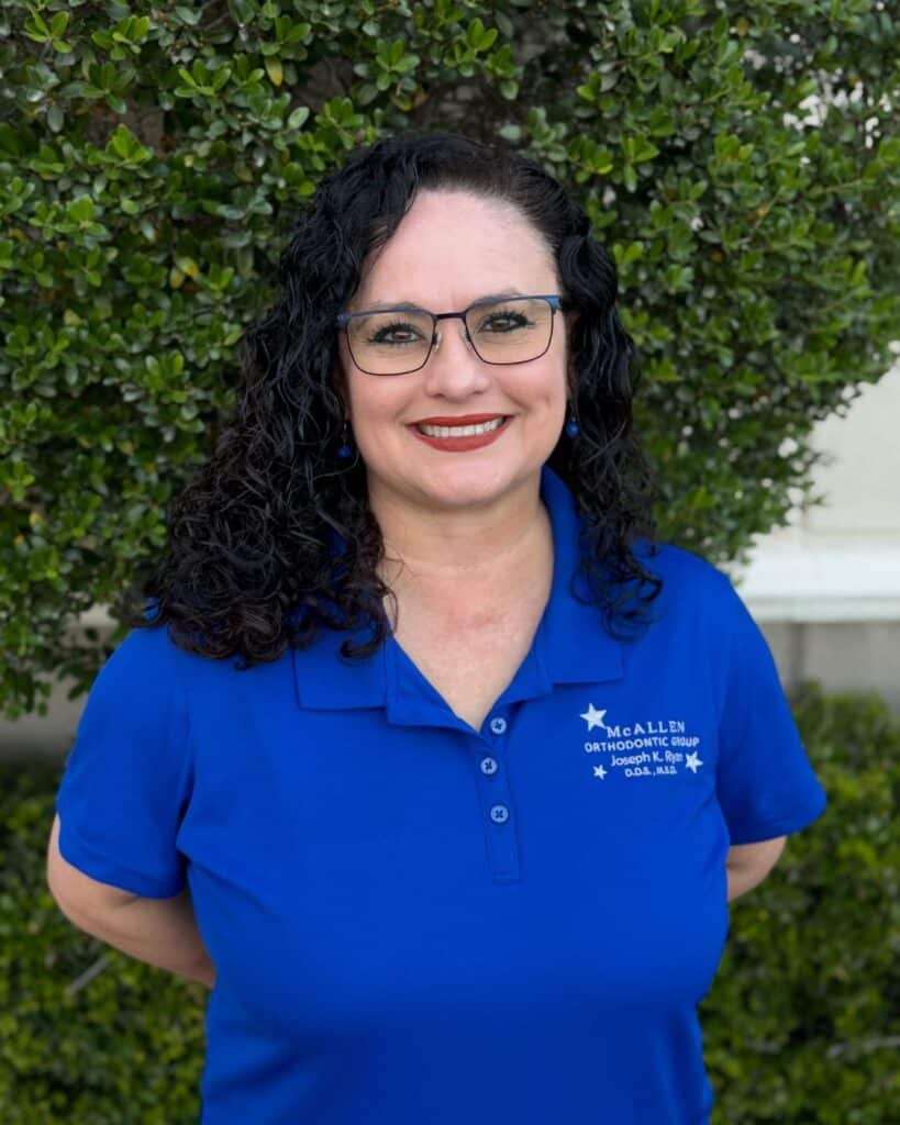 Portrait of Melissa Reyes, patient treatment manager at McAllen Orthodontic Group in McAllen, TX, wearing a blue clinic polo shirt and standing outdoors in front of greenery.